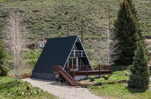 A modern A-frame cabin with a wooden deck, surrounded by greenery and hills.